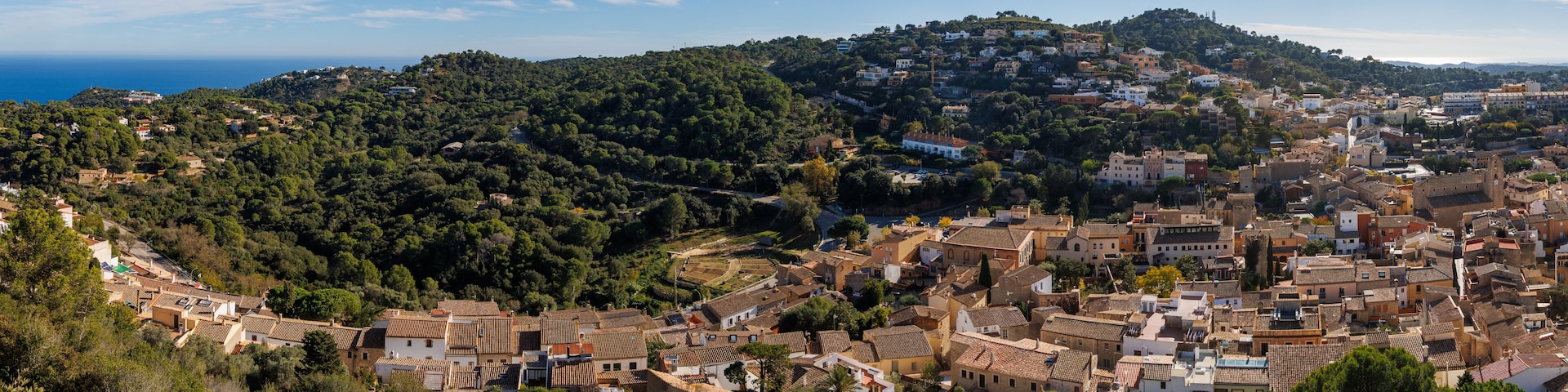 A coastal town with terracotta-roofed buildings, a hillside with dense greenery, and a clear view of the sea.