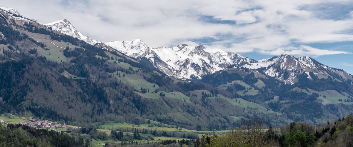 A mountainous landscape with snow-capped peaks, a valley with scattered houses, and a mix of evergreen and deciduous trees.