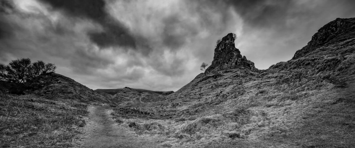 A rugged landscape with a narrow path, rocky outcrops, and sparse vegetation under a cloudy sky.
