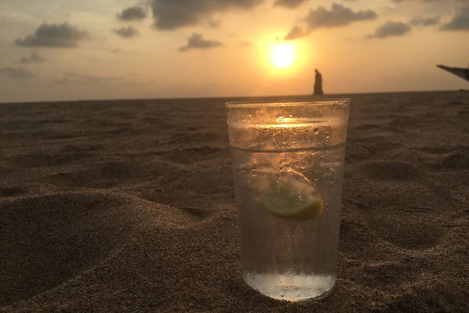 A glass of water with a lime slice on a sandy beach at sunset.