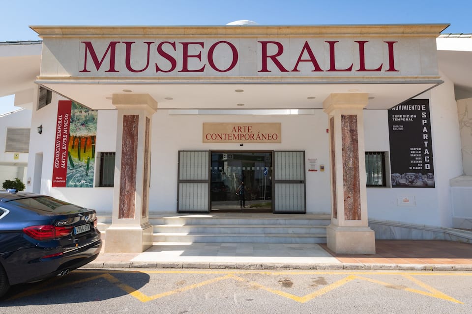 The entrance to Museo Ralli with a clear sign, a car parked in front, and a staircase leading up to the entrance.