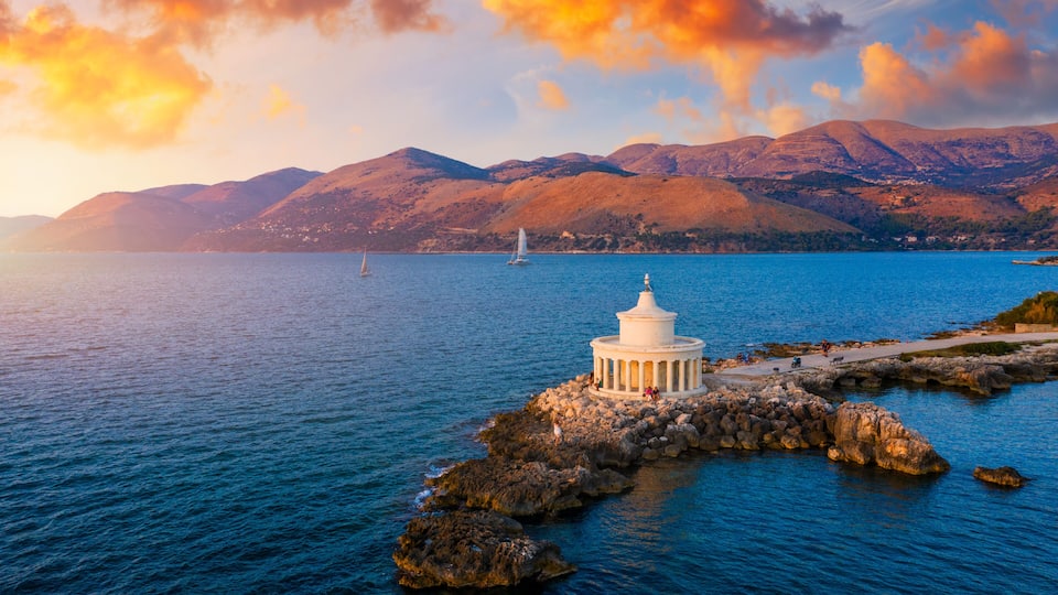 A coastal scene with a white lighthouse on a rocky outcrop, calm blue sea, and mountains in the background.