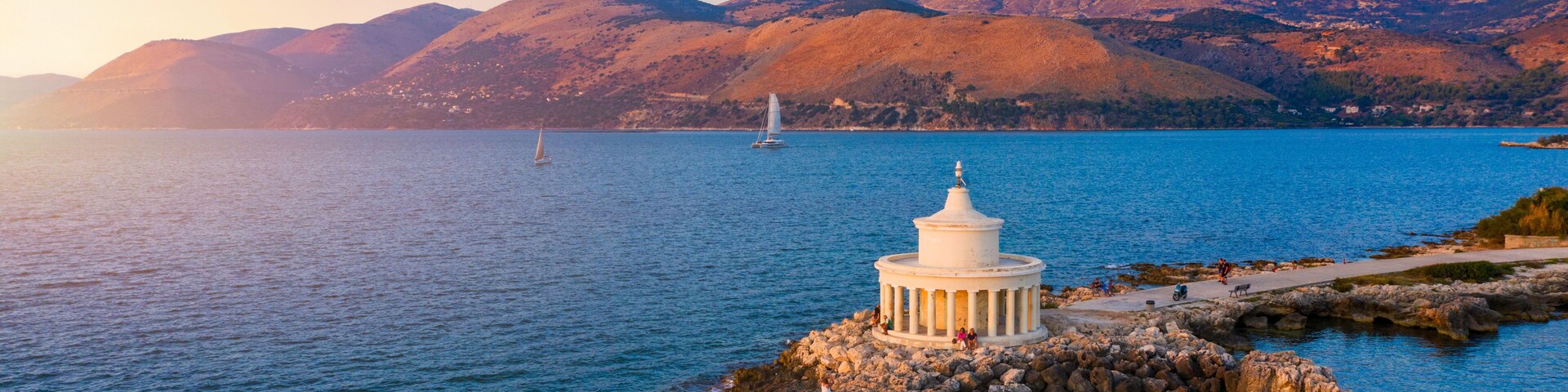 A coastal scene with a white lighthouse on a rocky outcrop, calm blue sea, and mountains in the background.