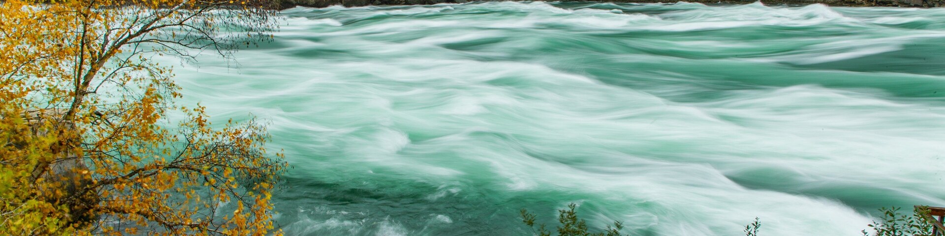Une rivière bordée d’une rambarde, des feuilles d’automne et une falaise rocheuse.