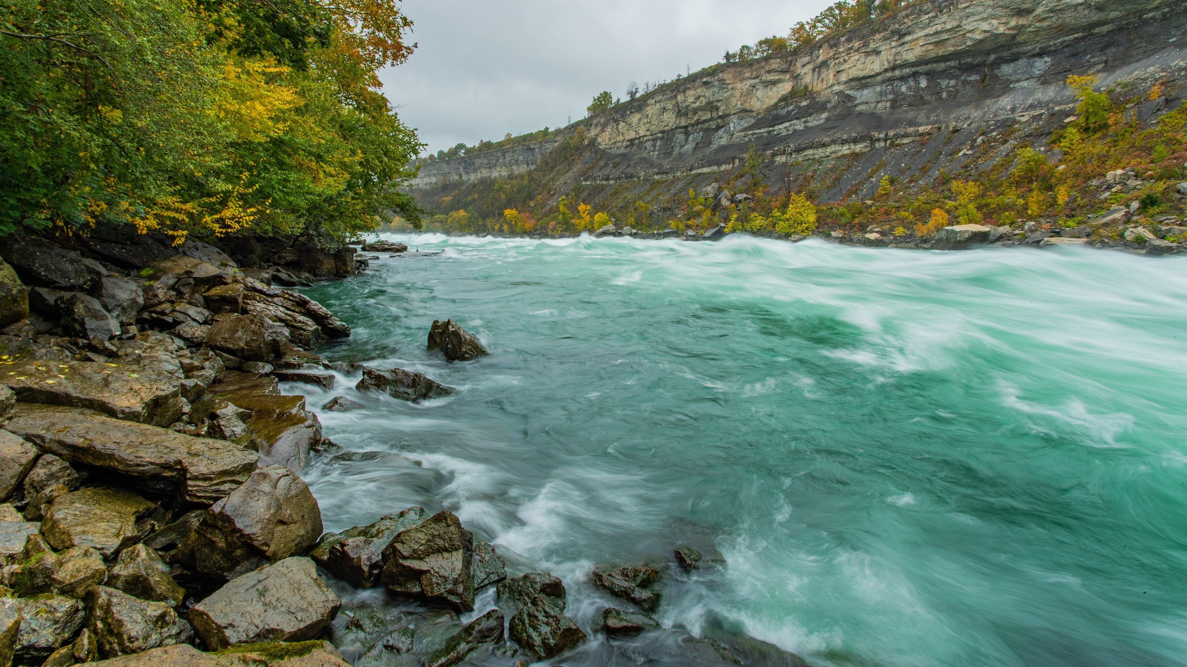 Ein Fluss mit schnell fließendem Wasser, umgeben von felsigem Gelände und Bäumen in Herbstfarben.