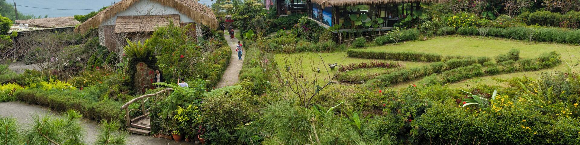 A lush garden with thatched-roof structures, terraced fields, and a pathway leading through the greenery.