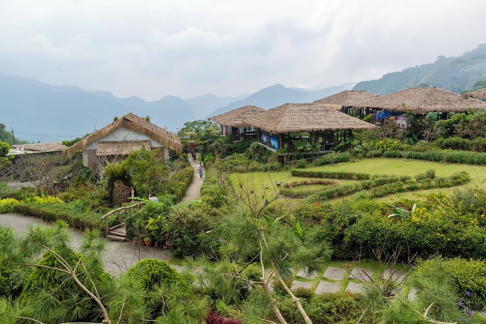 A lush garden with thatched-roof structures, terraced fields, and a pathway leading through the greenery.