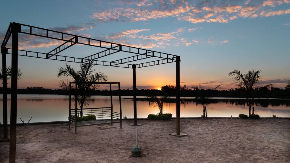 A metal structure with a bench, palm trees, and a calm lake at sunset.