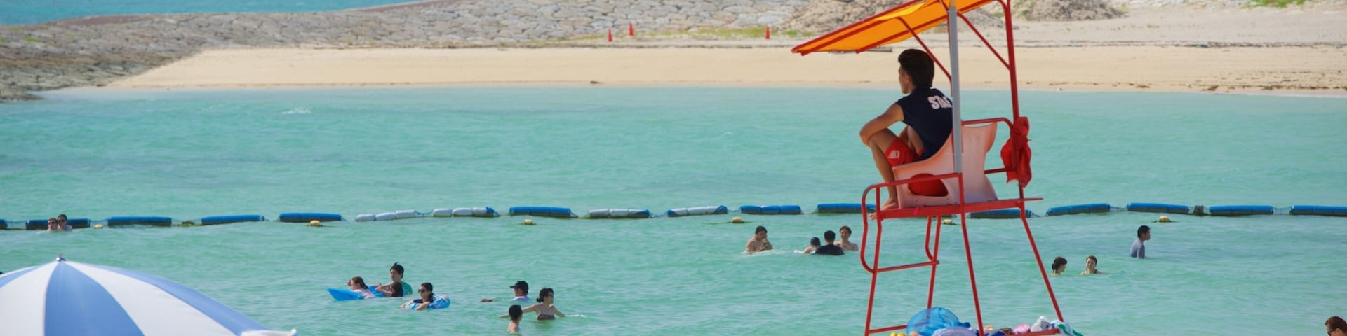 A lifeguard tower with a yellow flag, beach umbrellas, and people swimming in the ocean.