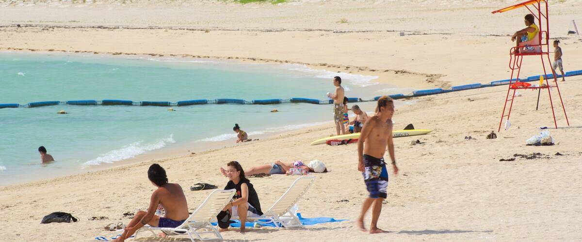 A sandy beach with people sunbathing, a lifeguard tower, and a few beach huts in the background.