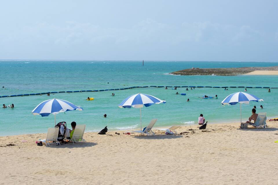 Una platja amb aigua turquesa cristal·lina, gent nedant i prenent el sol sota soledats de tires, i una roca al capvespre.