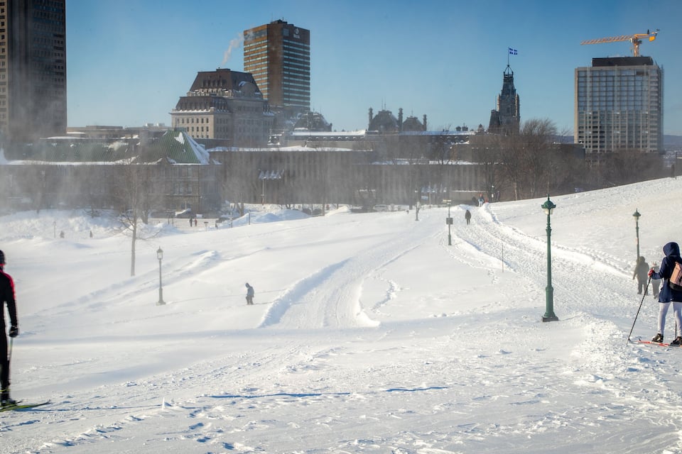 Una pista de esquí nevada con esquiadores, un horizonte urbano y un edificio histórico al fondo.