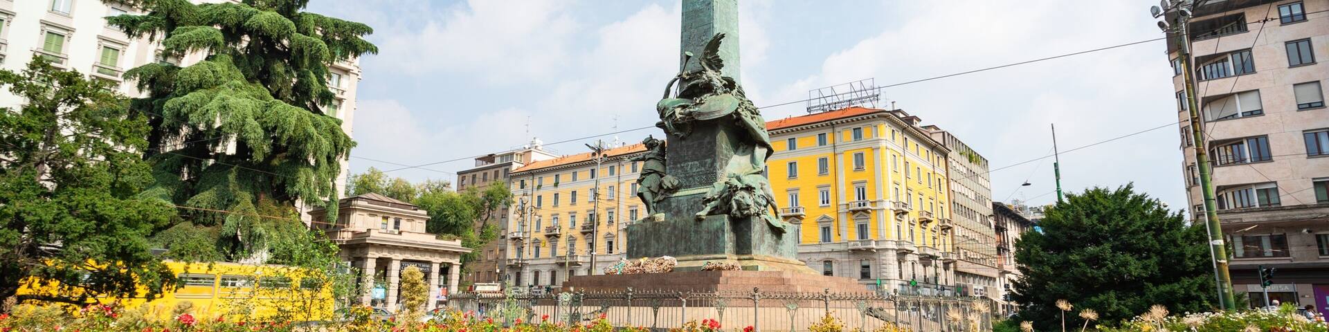 Un monumento con una alta obelisco, rodeado de flores coloridas y edificios de estilo clásico.