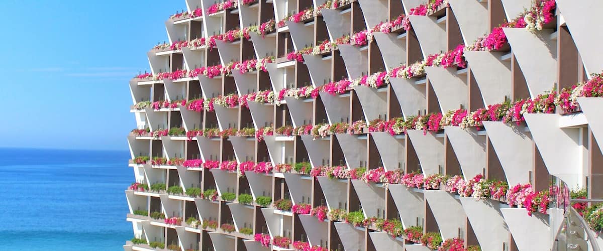 A modern hotel with balconies adorned with pink flowers, a clear blue sky, and a view of the ocean.
