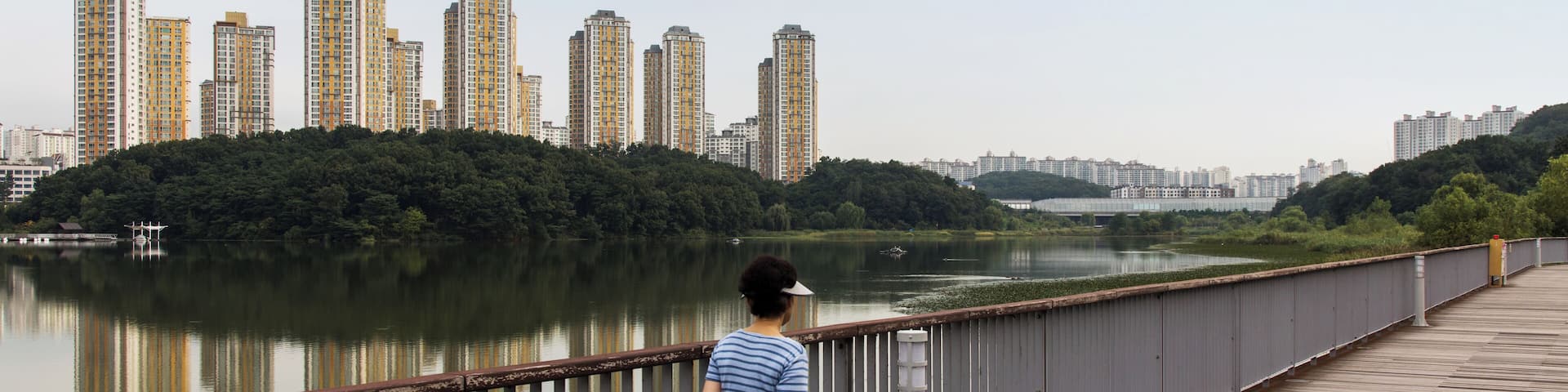 A woman standing on a wooden boardwalk overlooking a calm river with tall buildings in the background.