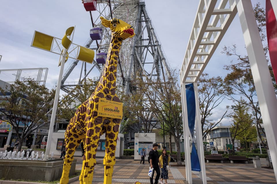 Una scultura di giraffa LEGO in una piazza pubblica, con una ruota panoramica sullo sfondo.