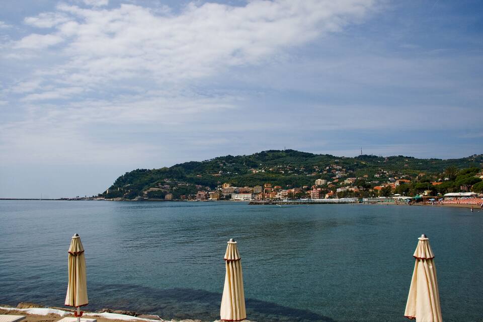 Vista sulla costa con acque limpide, ombrelloni sulla spiaggia e una cittadina collinare sullo sfondo.