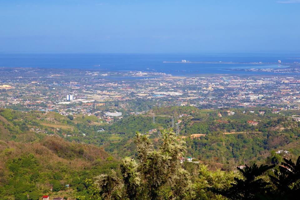 A coastal cityscape with dense urban development, green hills, and a clear view of the ocean.