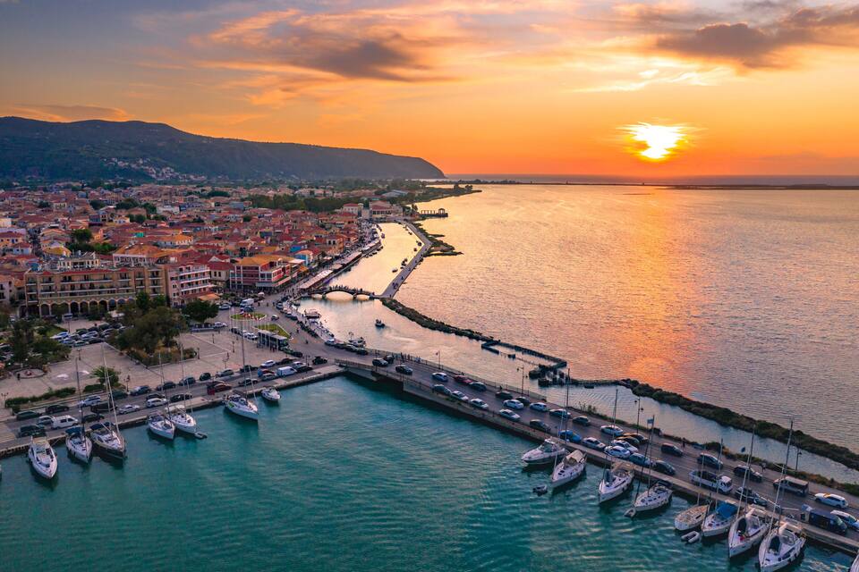 A coastal cityscape at sunset with a marina, boats, and a waterfront promenade.