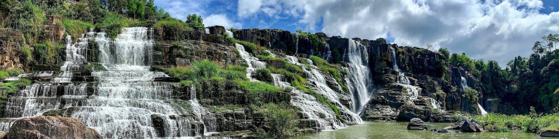 A multi-tiered waterfall cascading into a calm river surrounded by lush greenery and rocky cliffs.
