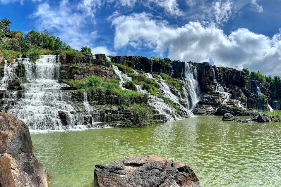Een meerlaagse waterval die in een rustige rivier stroomt, omringd door weelderige begroeiing en rotsachtige kliffen.