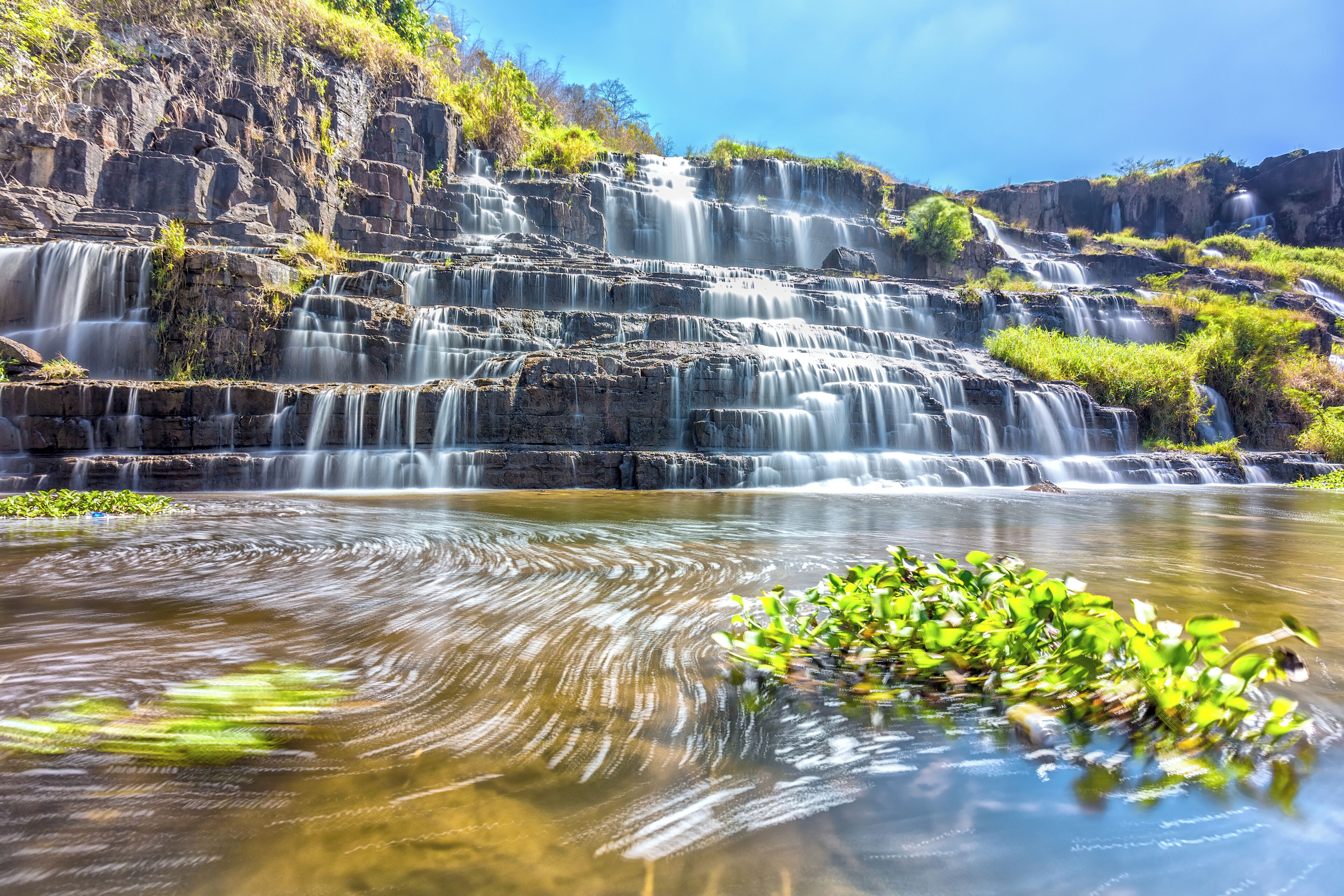 A multi-tiered waterfall cascading over rocky cliffs into a calm pool with green aquatic plants.