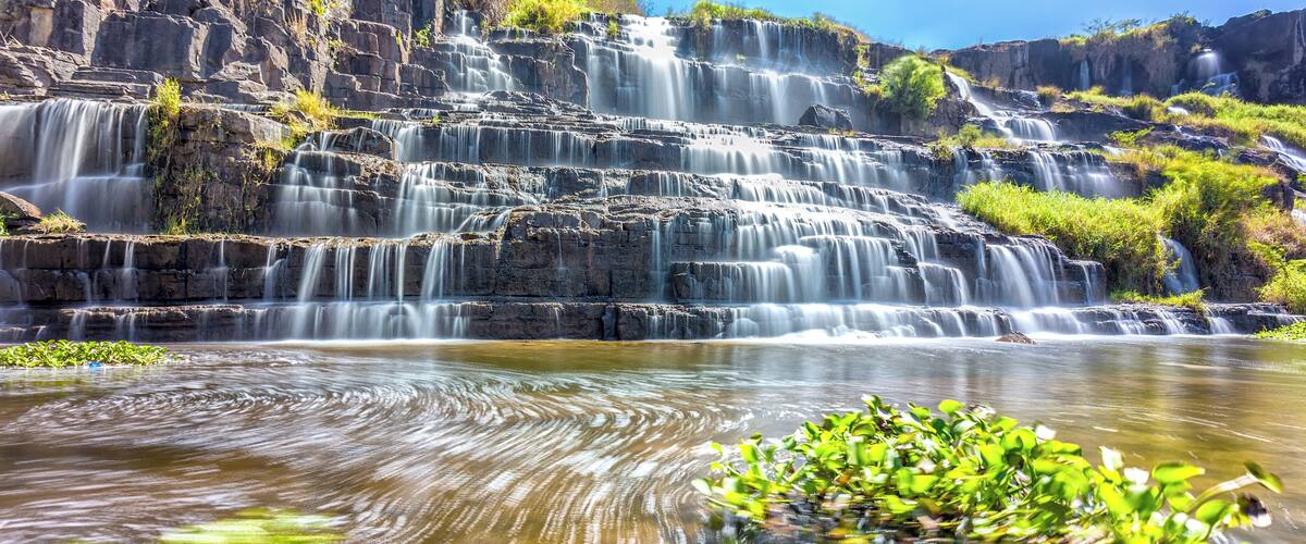 A multi-tiered waterfall cascading over rocky cliffs into a calm pool with green aquatic plants.