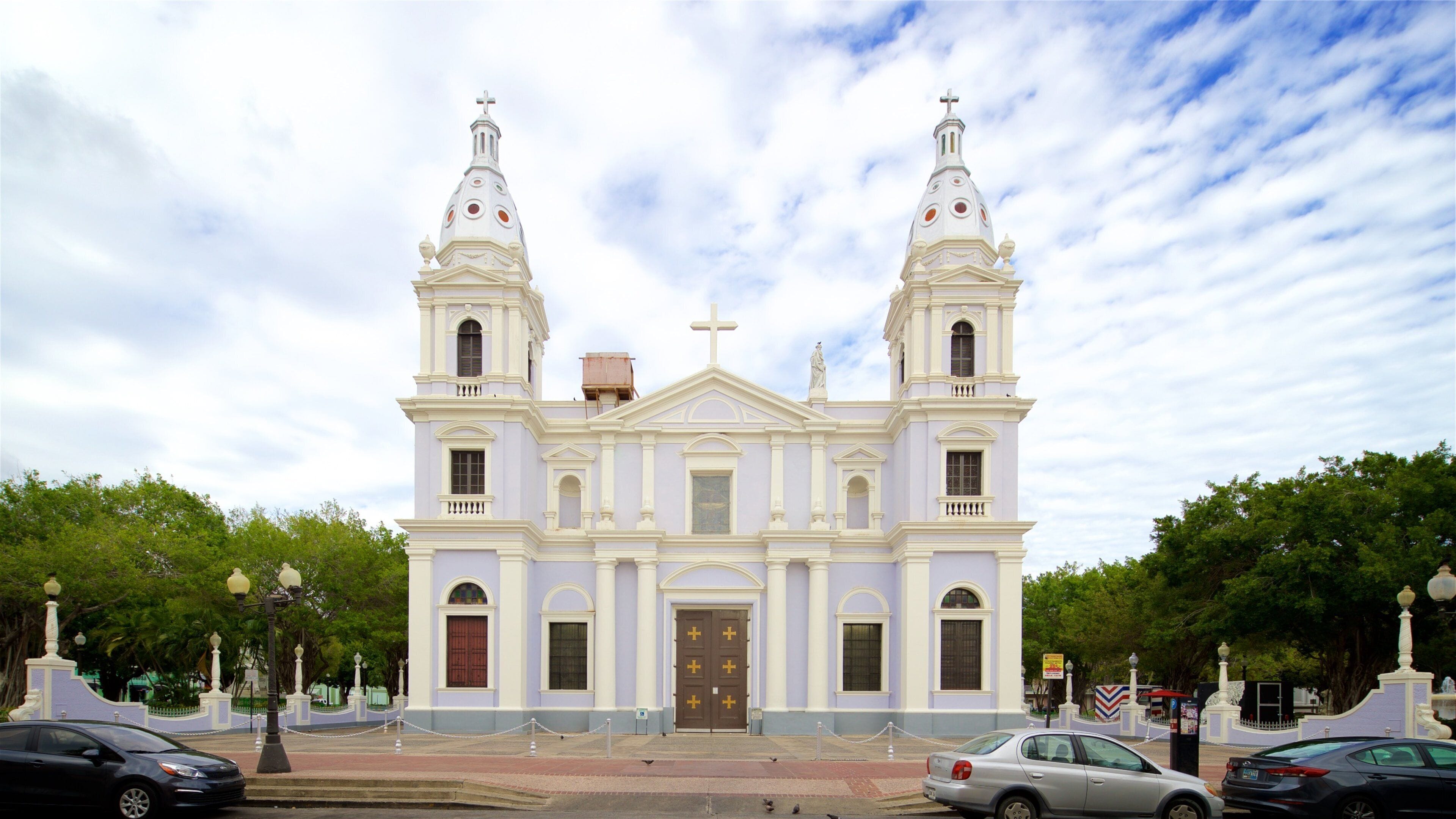 A white church with two towers and a central dome, surrounded by trees and parked cars.