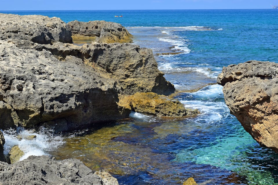 A rocky coastline with clear blue waters and waves crashing against the rocks.