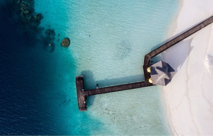 An aerial view of a beach with clear turquoise waters, a wooden pier, and a white sandy shore.