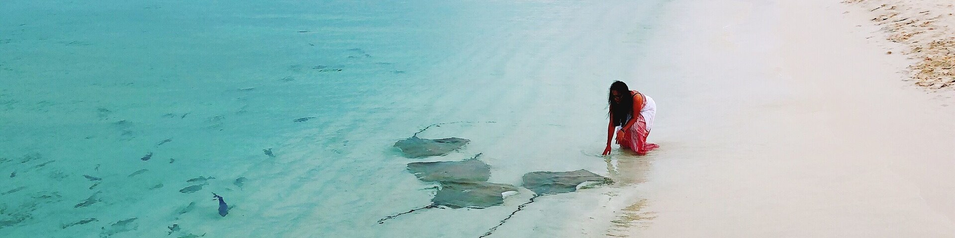 A beach with clear turquoise water and a person in a red dress walking along the shore.