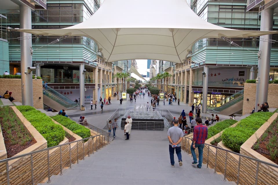En modern stadstorg med en central fontän, omgiven av grönska och med rulltrappor som leder upp till de övre våningarna.