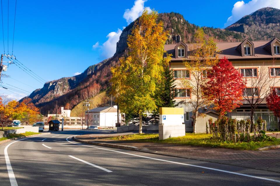 A road curving through a residential area with autumn-colored trees, a mountain in the background, and a multi-story building with a parking sign.