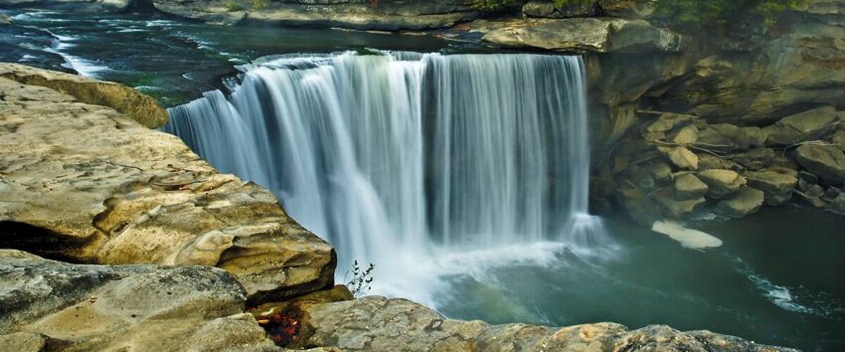 A waterfall cascading over rocks into a river surrounded by forested hills.
