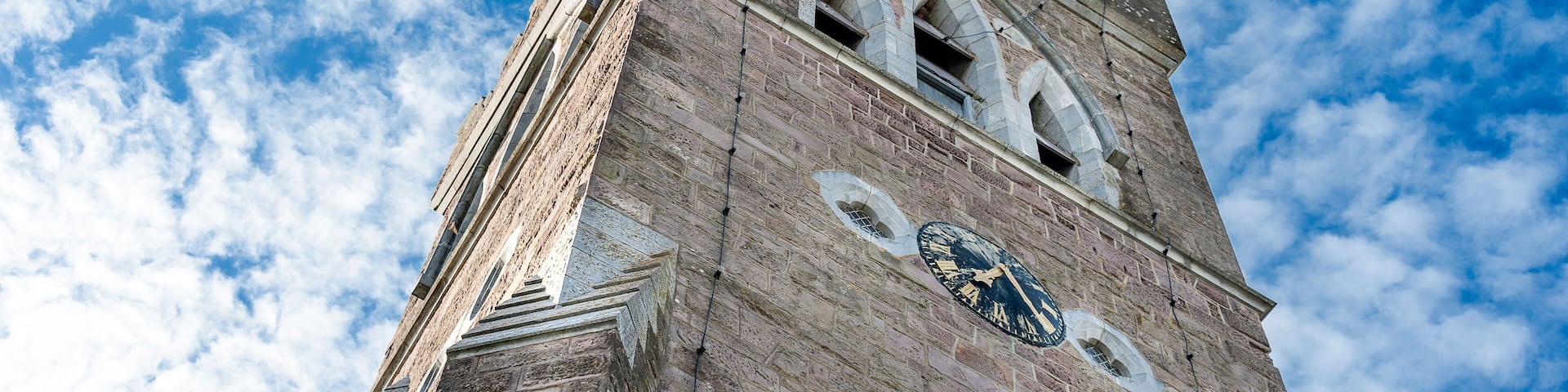 Une haute tour en briques, surmontée d’une horloge et dotée de fenêtres en arc de cercle, se dressant sur un ciel bleu parsemé de nuages.
