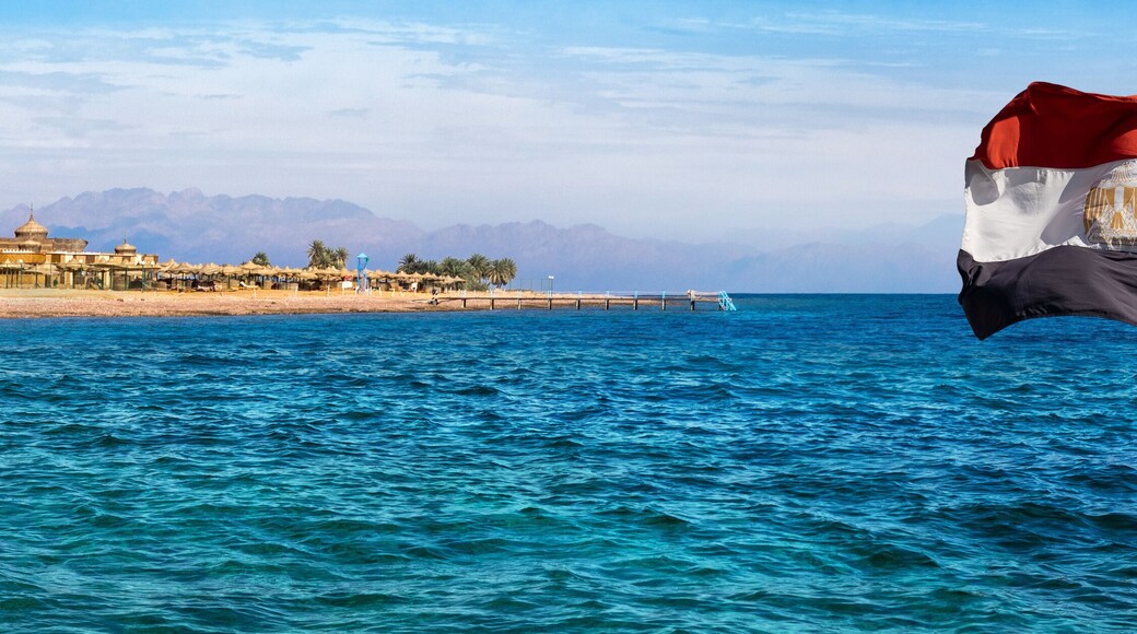 Aqaba bay view near the Dahab resort with a hotel, palms and silhouettes of mountains at skyline. Panorama of the coastline of Red Sea with Egyptian flag on a beautiful beach in Egypt.
