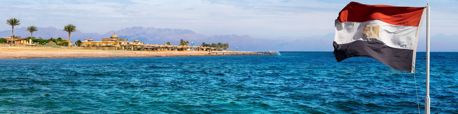 Aqaba bay view near the Dahab resort with a hotel, palms and silhouettes of mountains at skyline. Panorama of the coastline of Red Sea with Egyptian flag on a beautiful beach in Egypt.
