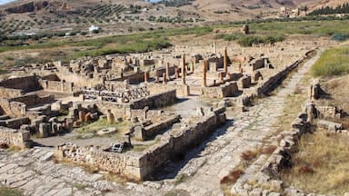 Ancient ruins with stone pathways and columns, set against a backdrop of hills and sparse vegetation.