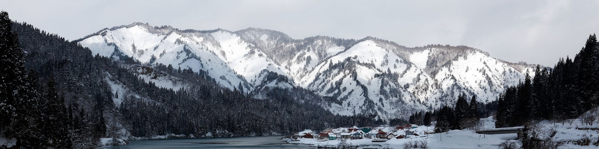 Beautiful scenery of snowy mountains reflected in the peaceful water of River Tadami Gawa and a village perched on the riverside slope on a sunny, cloudy winter day in Oku-Aizu, Fukushima, Japan