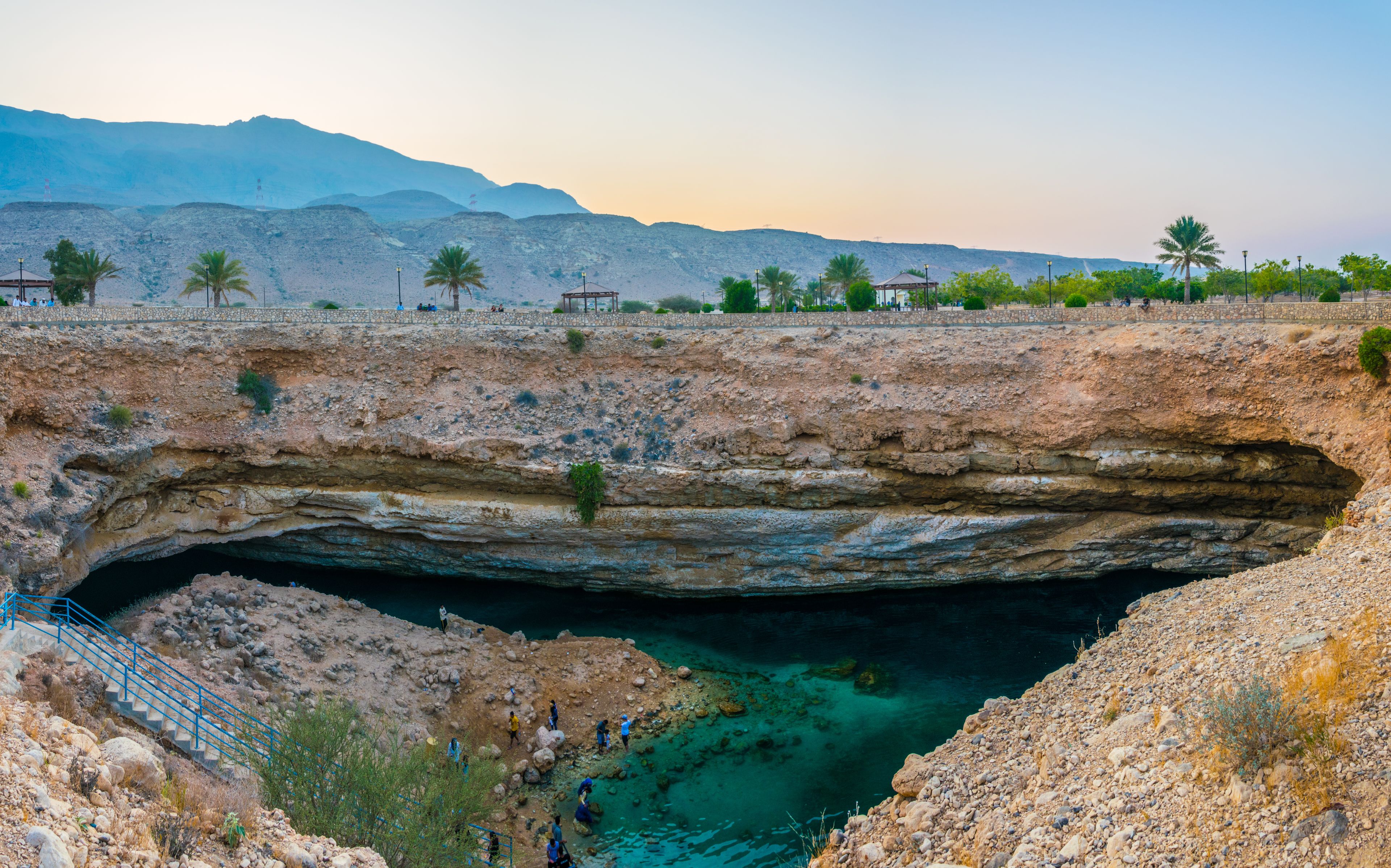 A natural rock formation with a cave entrance, palm trees, and distant mountains.