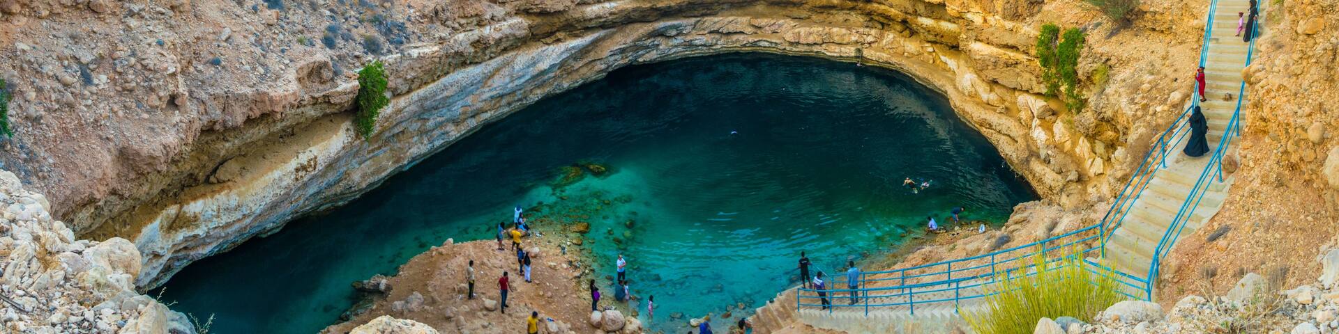 A large natural sinkhole with a clear blue pool at its bottom, surrounded by rocky terrain and a few people walking around.