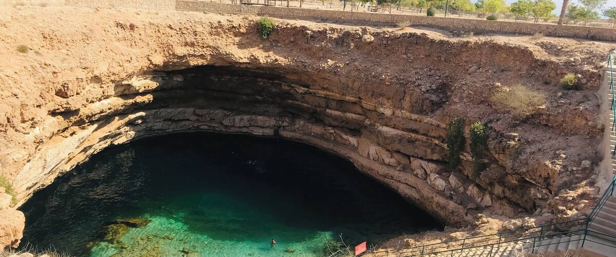 A natural cave opening with clear water, surrounded by rocky terrain and sparse vegetation.