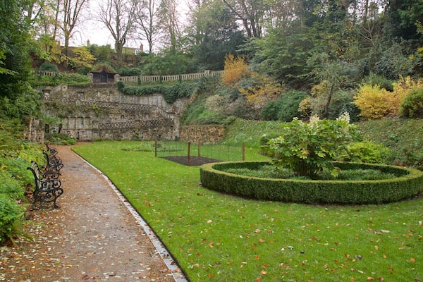 A garden with a stone wall, a circular hedge, and a pathway with benches.