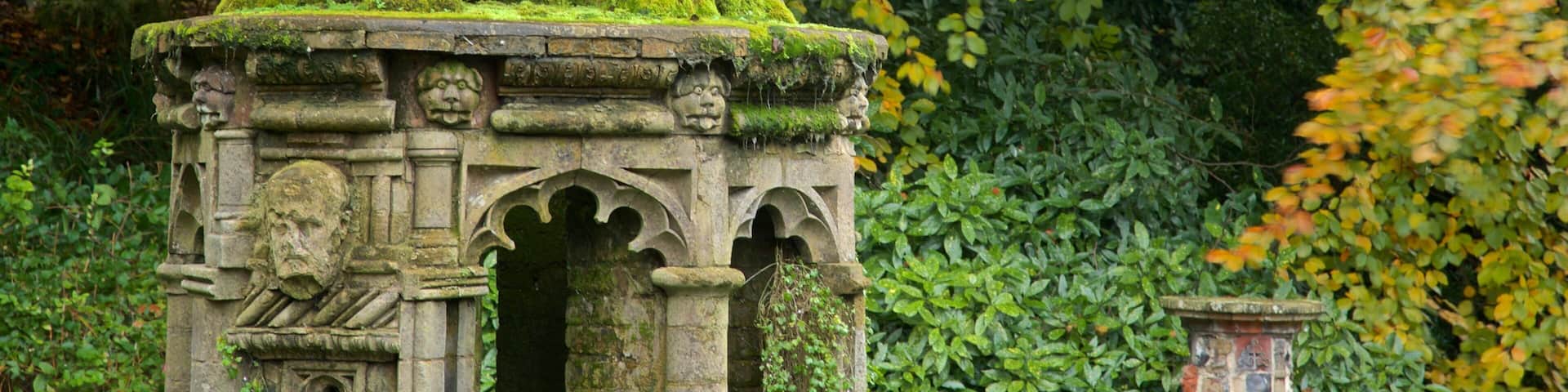 Una torre de piedra con columnas y arcos cubiertos de musgo, rodeada de vegetación exuberante y hojas otoñales.