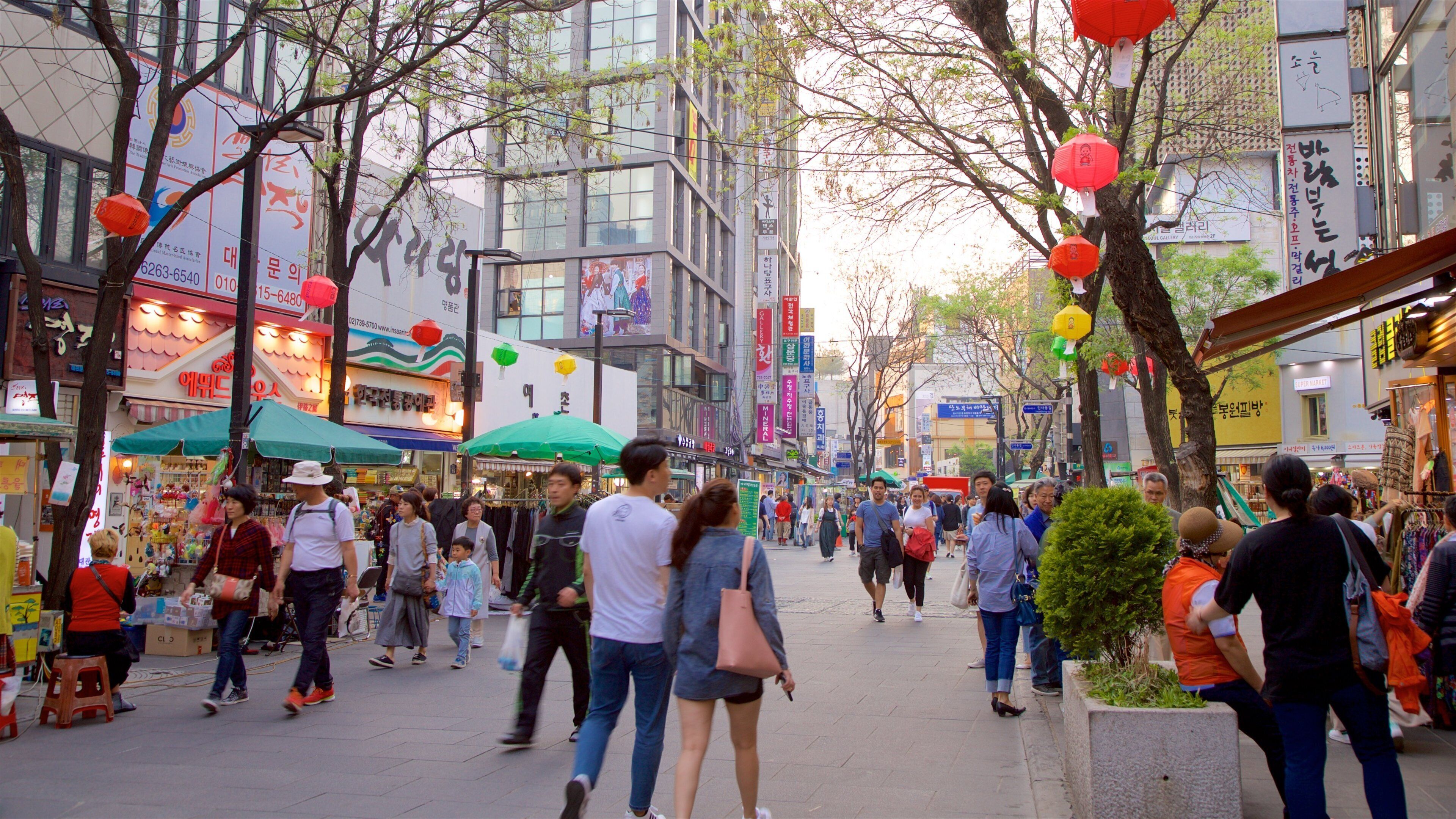 A bustling urban street lined with shops and trees, with people walking and carrying shopping bags.