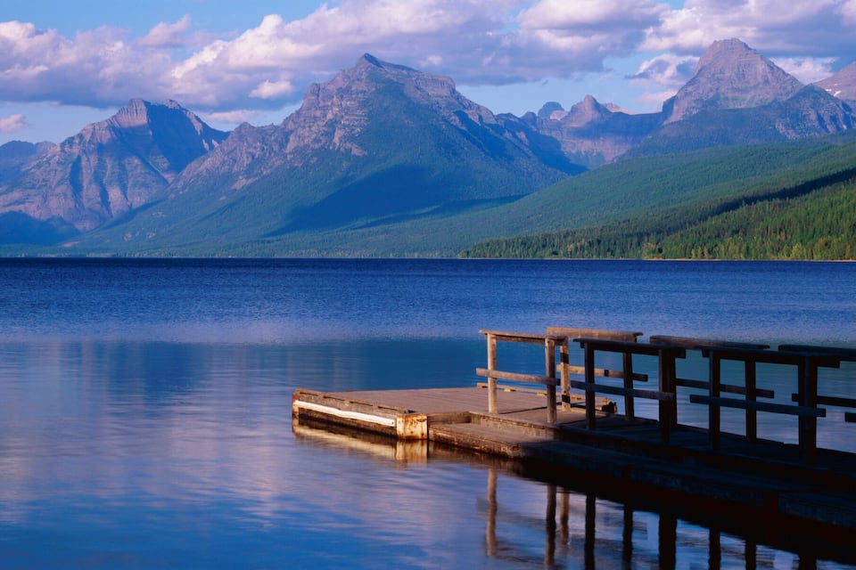 Un lago tranquillo con un molo di legno che si protende nell'acqua, circondato da montagne boscose sotto un cielo parzialmente nuvoloso.