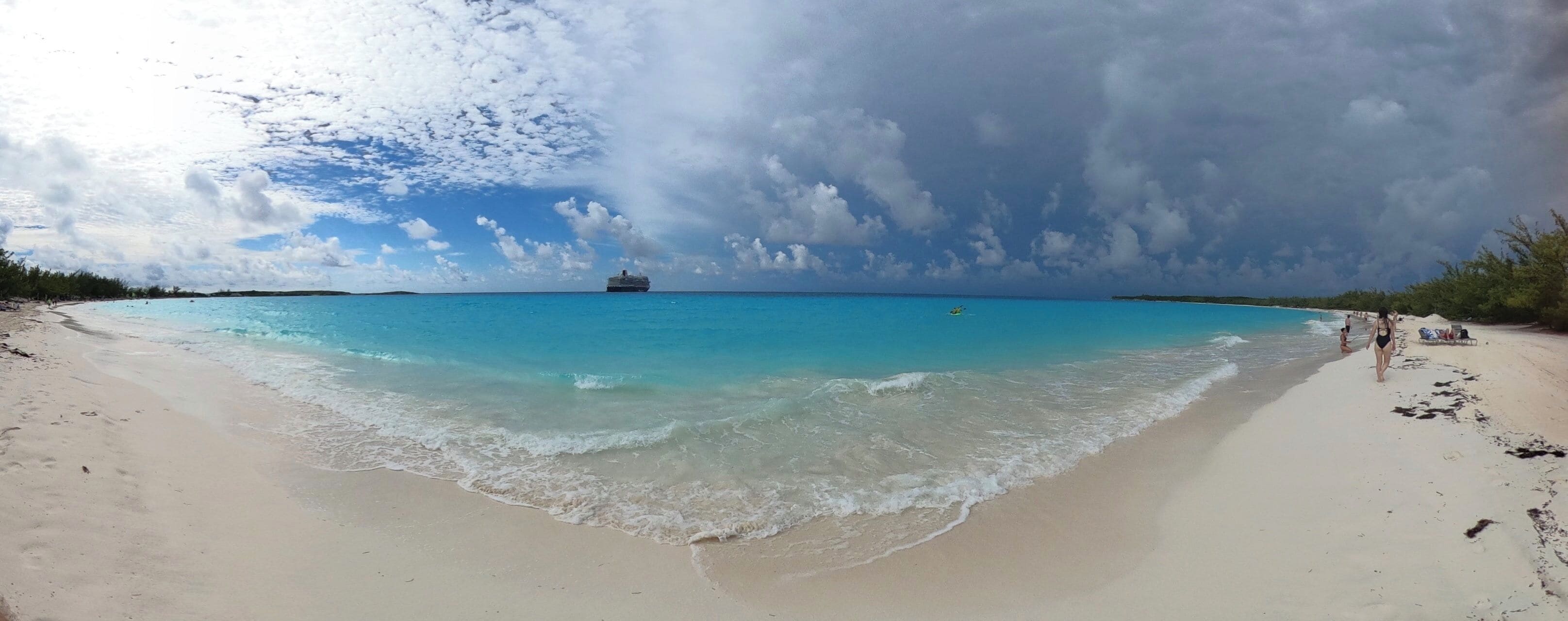 A beach with clear turquoise water, a cruise ship in the distance, and a partly cloudy sky.