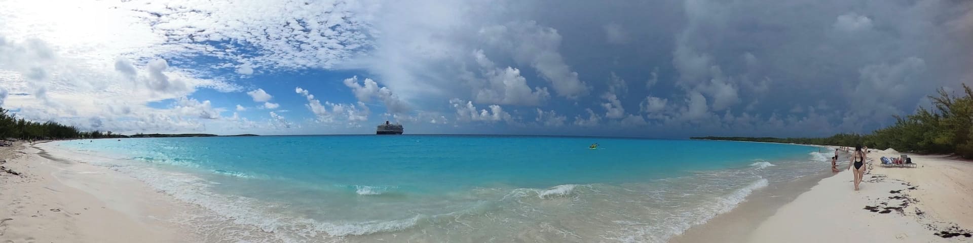 A beach with clear turquoise water, a cruise ship in the distance, and a partly cloudy sky.