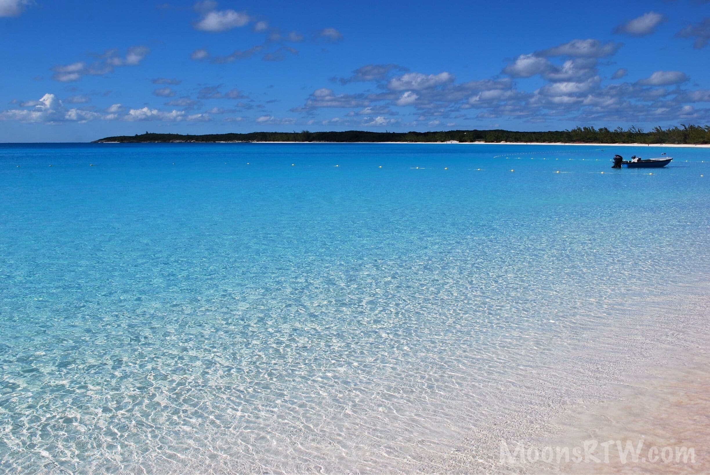 A clear blue sea with a boat on the right, a sandy beach, and a distant shoreline with trees.