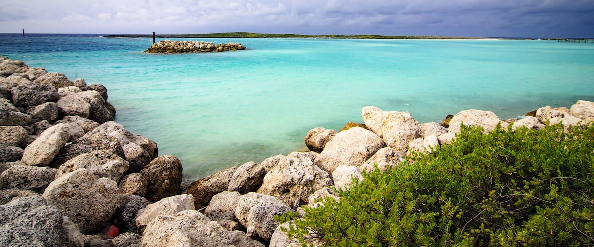 A rocky shoreline with clear turquoise water, a distant pier, and overcast sky.
